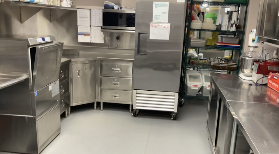 Commercial kitchen with a stainless steel refrigerator, prep counter, and shelving stocked with supplies