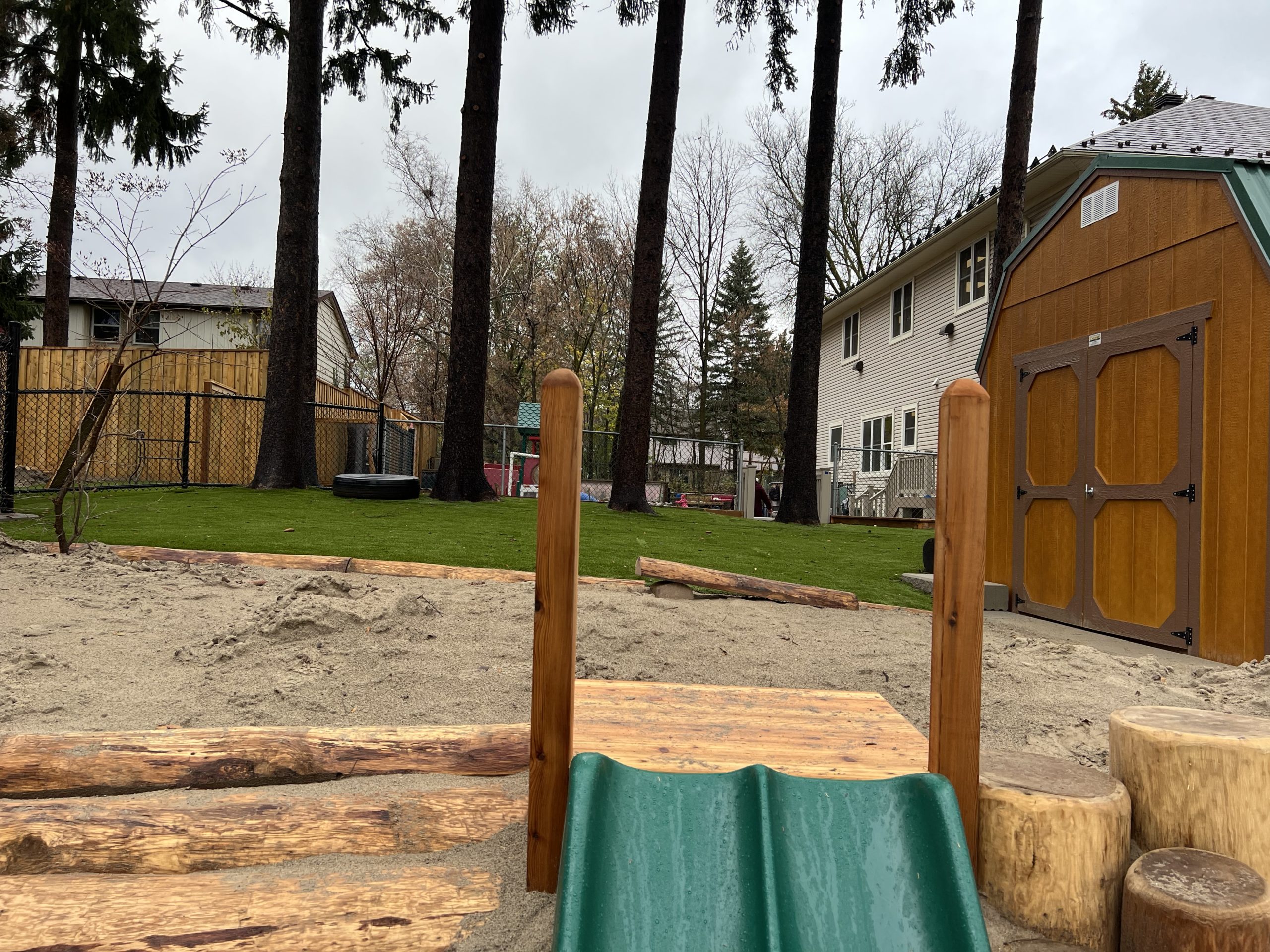 Sand pit connected to a grassy turf lawn shaded by mature trees with a storage shed