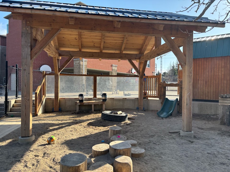 Sand play area under the pavilion with a tire sandbox, tree-stump stepping stones, a small slide, and a bench