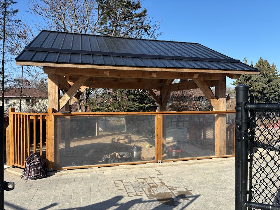 Covered wooden pavilion with a black metal roof over a sand play area, enclosed by a mesh and wood fence