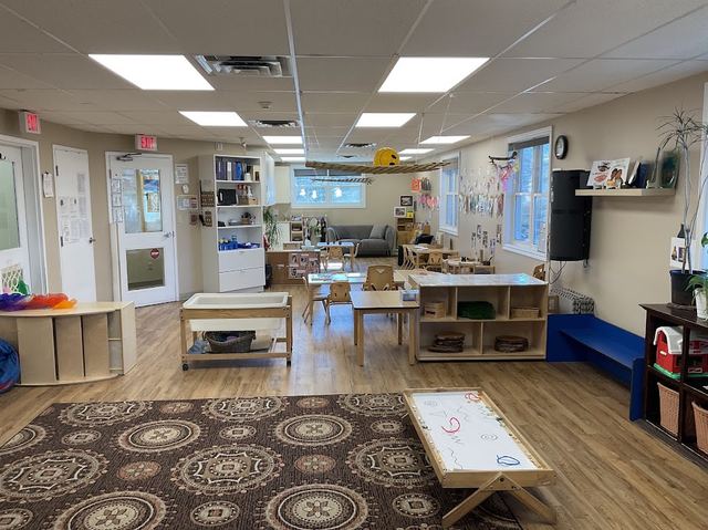 Wide classroom view showing an area rug, group tables, and open shelving