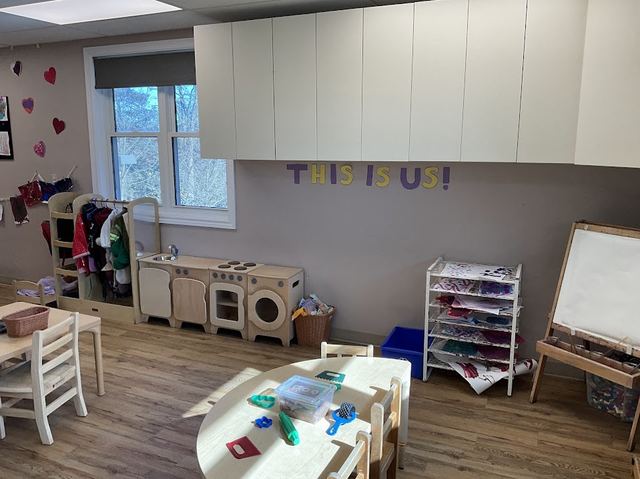 A classroom wall reading This Is Us above a dress-up clothing rack and a wooden play kitchen set