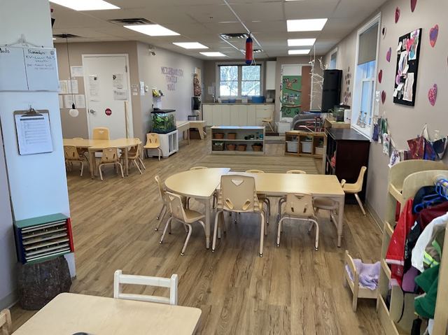 A preschool classroom with tables and chairs, a Neighbourhood Family Wall mural, a fish tank, and a coat rack decorated with children's heart artwork