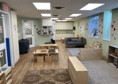 A classroom featuring a patterned area rug, gray couch, bookshelf, activity table, and a staff kitchenette counter in the background, decorated with paper hearts.