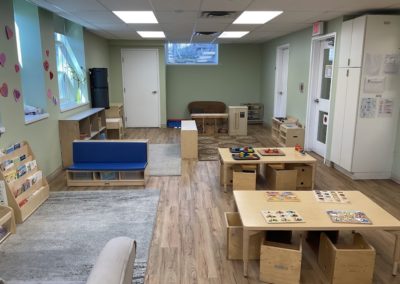 A room with gray rugs, a blue cushioned bench, wooden play furniture, activity tables with puzzles, and a bookshelf.