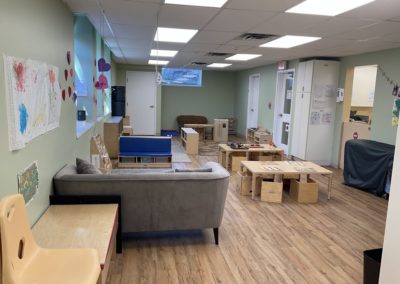 A long green-walled room with a gray couch, wooden play furniture, storage cubes, and several doors leading to other rooms.
