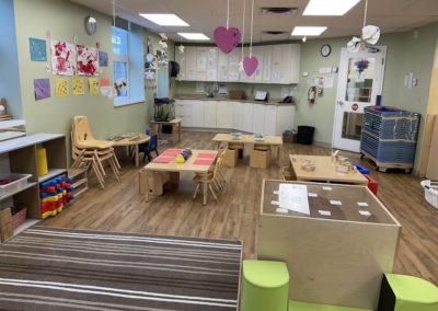 A wide view of a toddler classroom showing multiple activity tables, storage cubbies, a play cube, and hanging paper hearts under fluorescent lighting.