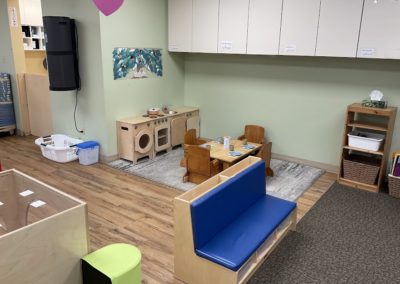 A wooden play kitchen set beside a small dining table and chairs on a gray rug, with a blue cushioned bench and storage bins nearby.