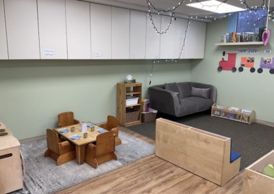 A small wooden table with chairs set for snack time, next to a gray loveseat, storage cubbies labeled with staff names, and string lights overhead.