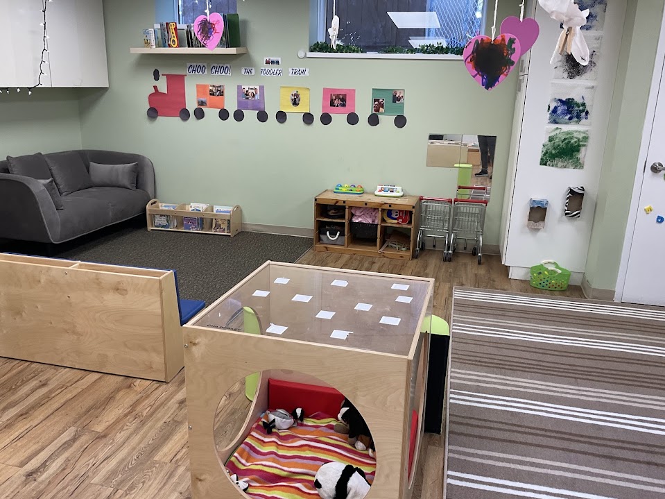 Cozy reading nook with a gray couch, bookshelf, and wooden play cube with stuffed animals in a toddler classroom