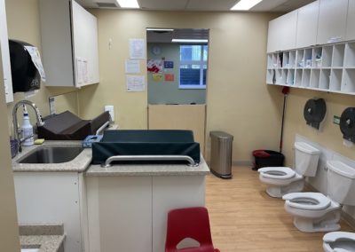 A daycare washroom with a diaper-changing counter, sink, paper towel dispenser, stainless steel trash can, and a row of three low child-height toilets along the wall.