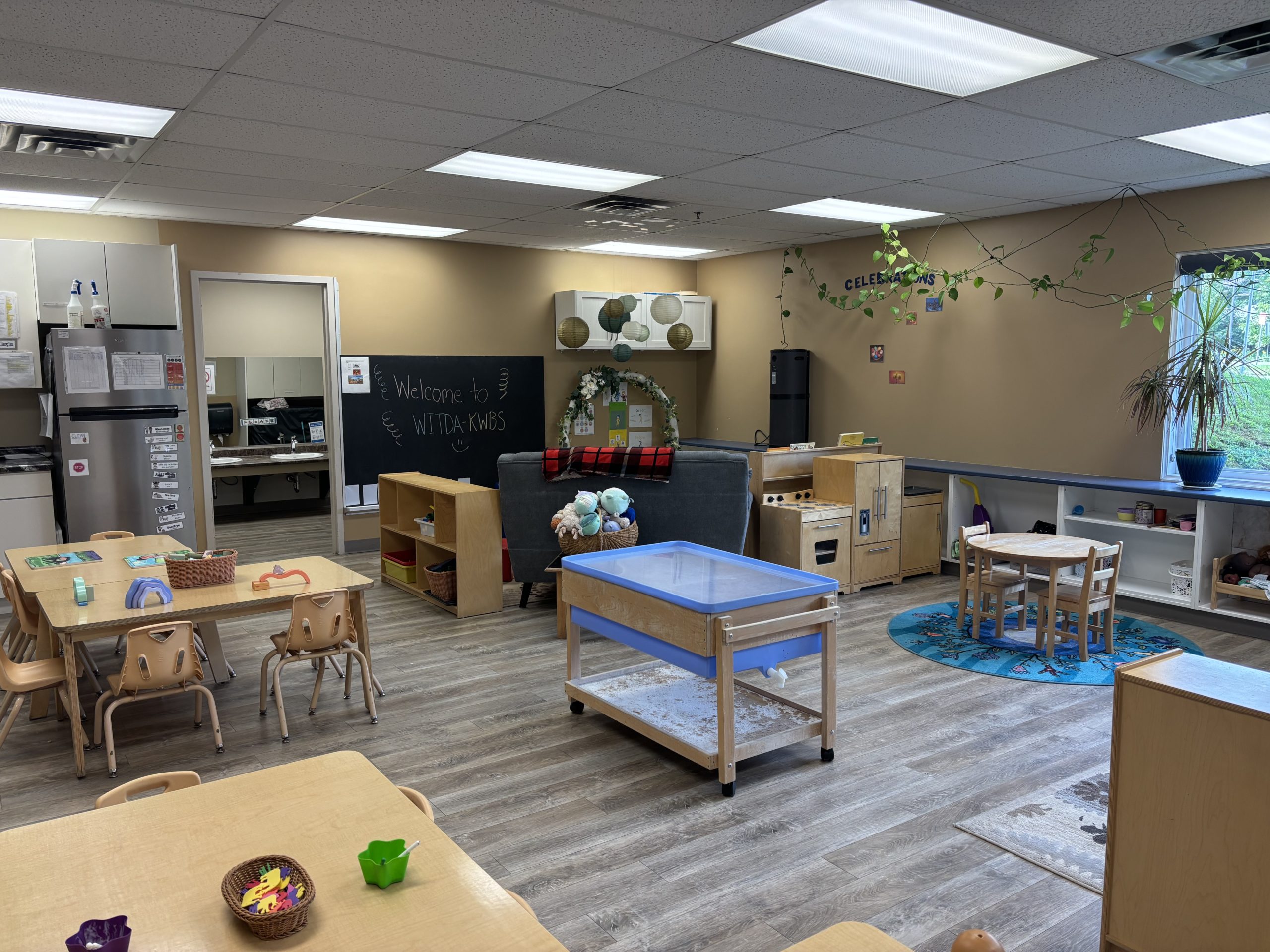 Classroom with a wooden play kitchen, small round table, welcome chalkboard and kitchenette