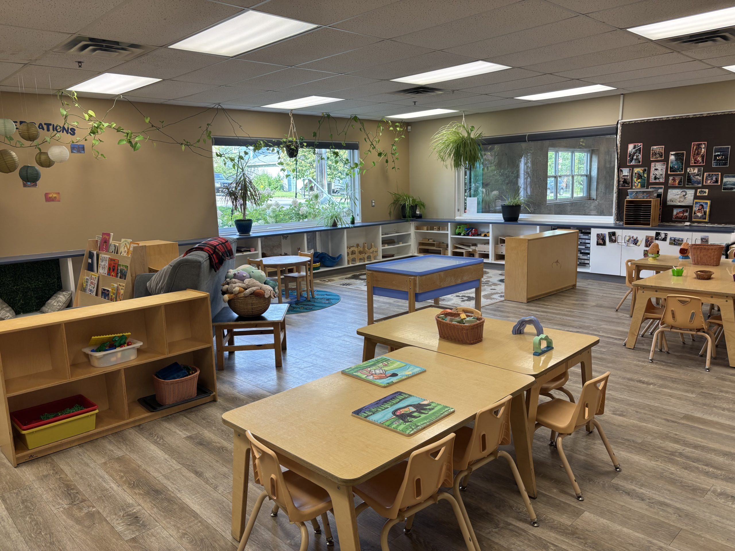Bright classroom with child-sized tables and chairs, a reading nook, sensory table and plants along the windows