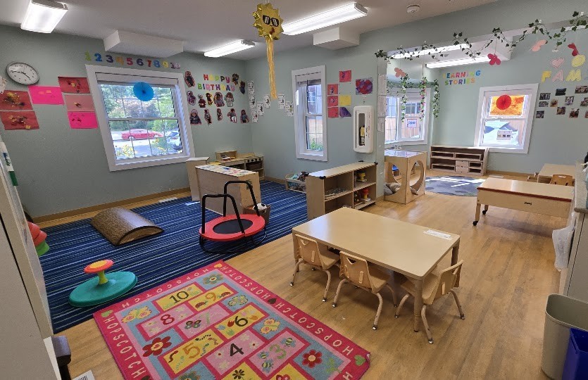 Play area with a blue striped carpet, a mini trampoline with a handle, low toy shelves and a birthday display on the wall